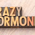 Eine Frau sitzt mitten im Alltag, lächelt nach außen und hält innerlich ein wild durcheinanderwirbelndes Hormon-Orchester aus Gedanken, Emotionen und Erschöpfung zusammen, auf der Suche nach ihrem Hormongleichgewicht.